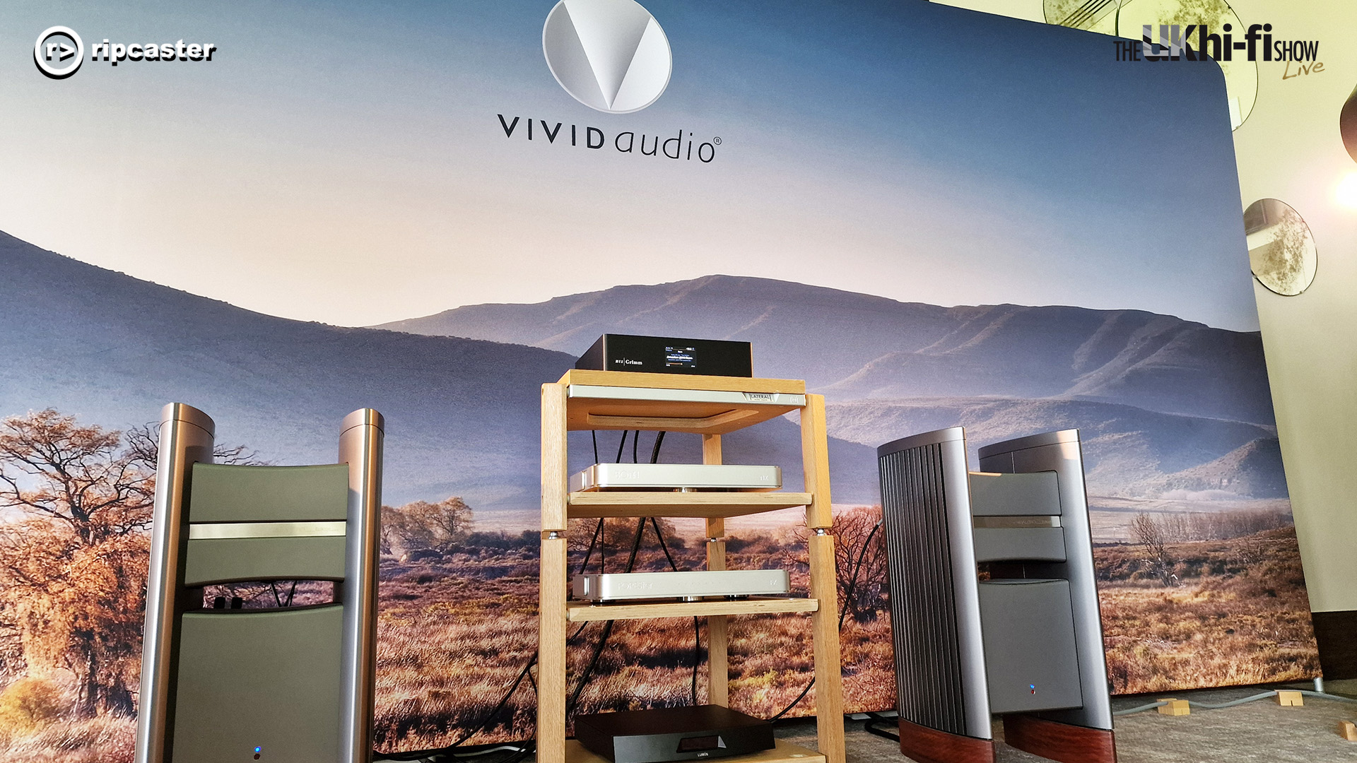 Vivid audio.  A pair of floorstanding speakers either side of some hifi equipment.  the backdrop says "vivid audio" and has a landscape pictured