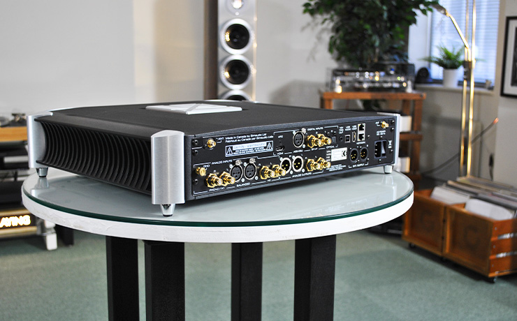 The Moon 791 on a table in the ripcaster showroom.  The view is of the back showing all the connections and an on/off switch.  In the background are large Audiovector floorstanding speakers and other pieces of audio kit.  there are two boxes of records on the floor.
