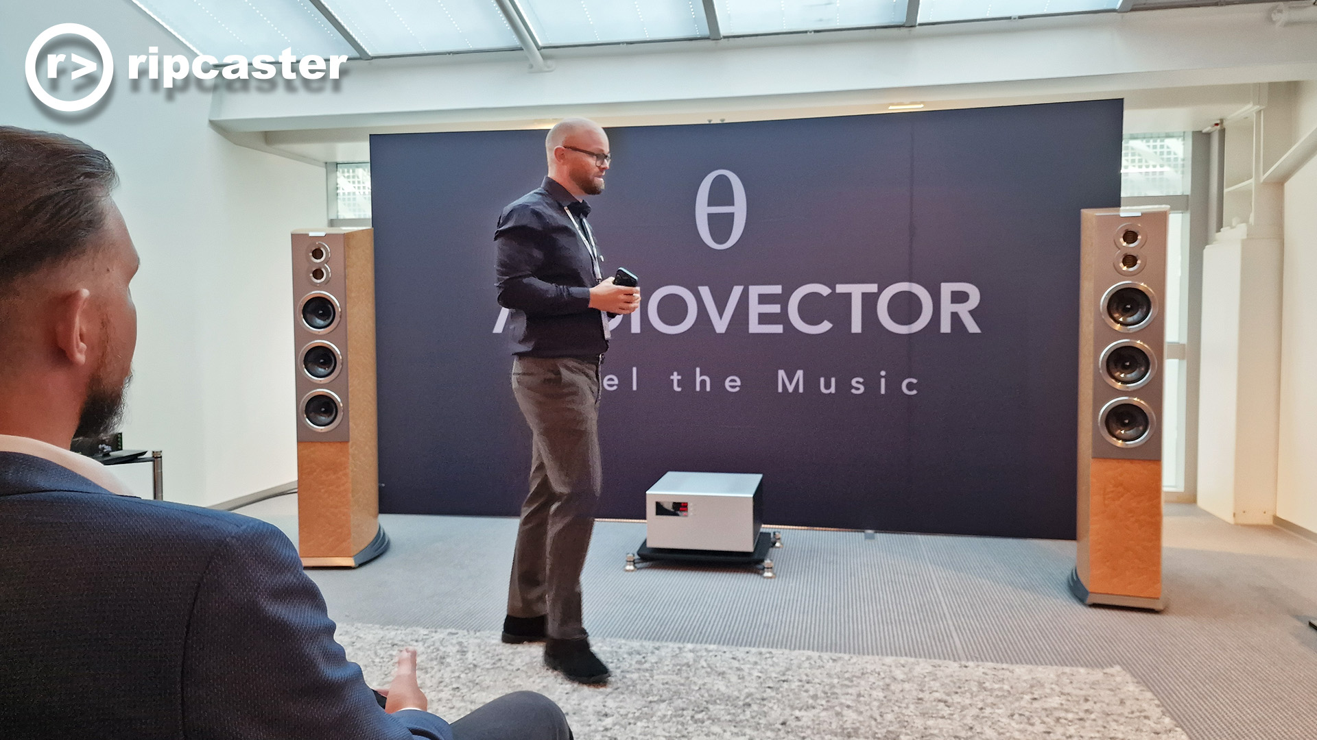 A man stands in front of a large backdrop that says "Audiovector, Feel the Music".  There are large floorstanding speakers either side of the sign and just in front of it.  There's a man partially visible in the foreground.