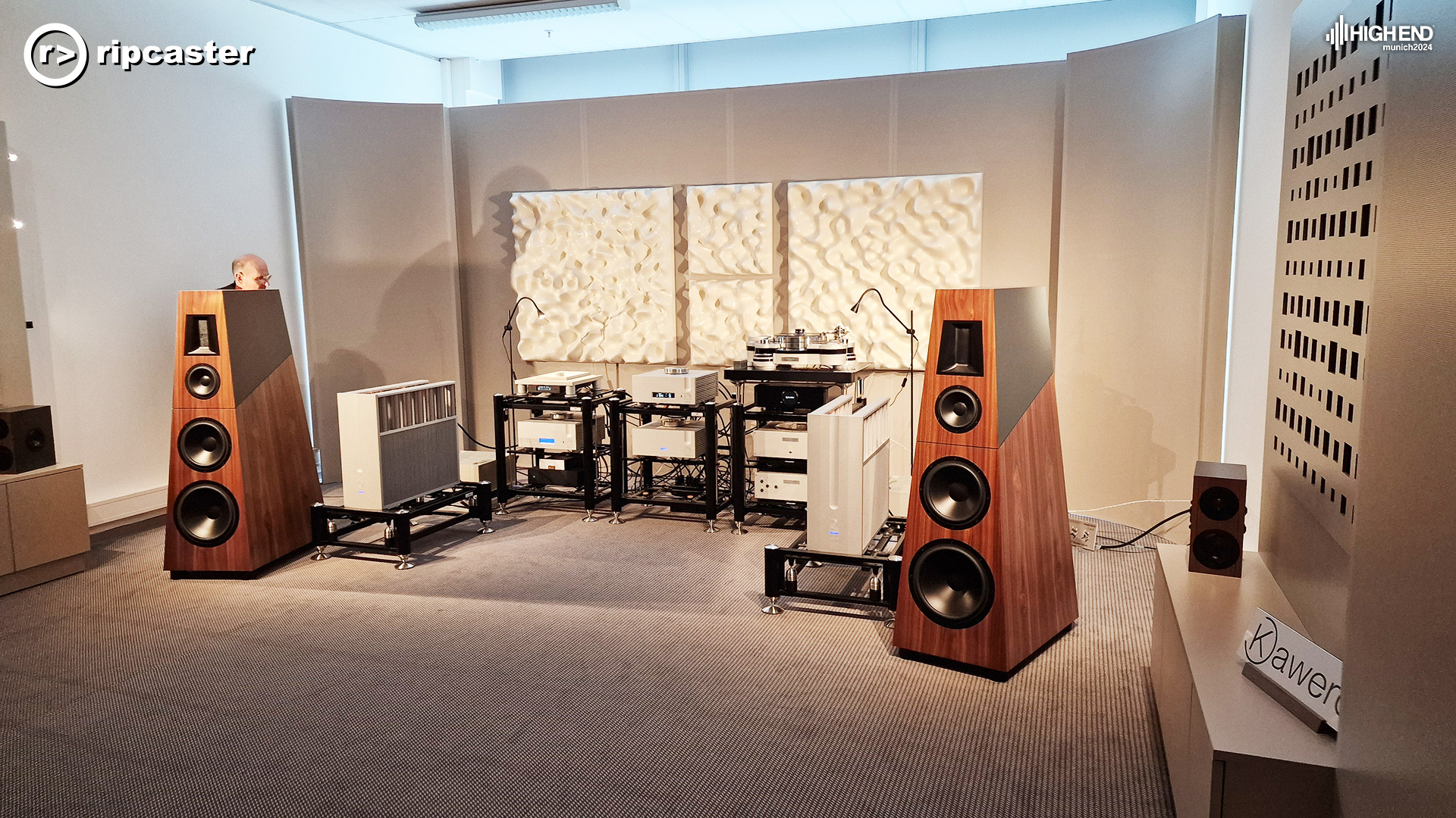 A pair of HiFi speakers either side of HiFi equipment.  The speakers are floorstanding and narrower at the top than the bottom.  there's a man's head just visible over the top of the left speaker.