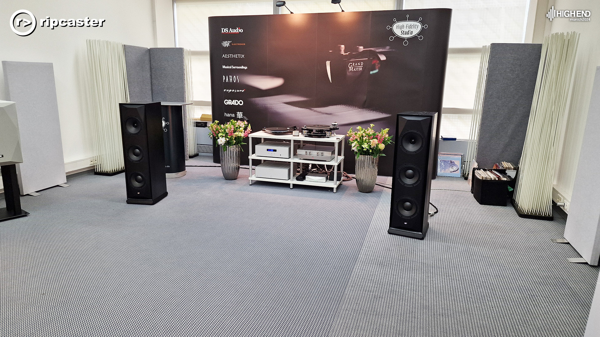 A pair of black floorstanding speakers in a sparse room with HiFi equipment and large vases of flowers