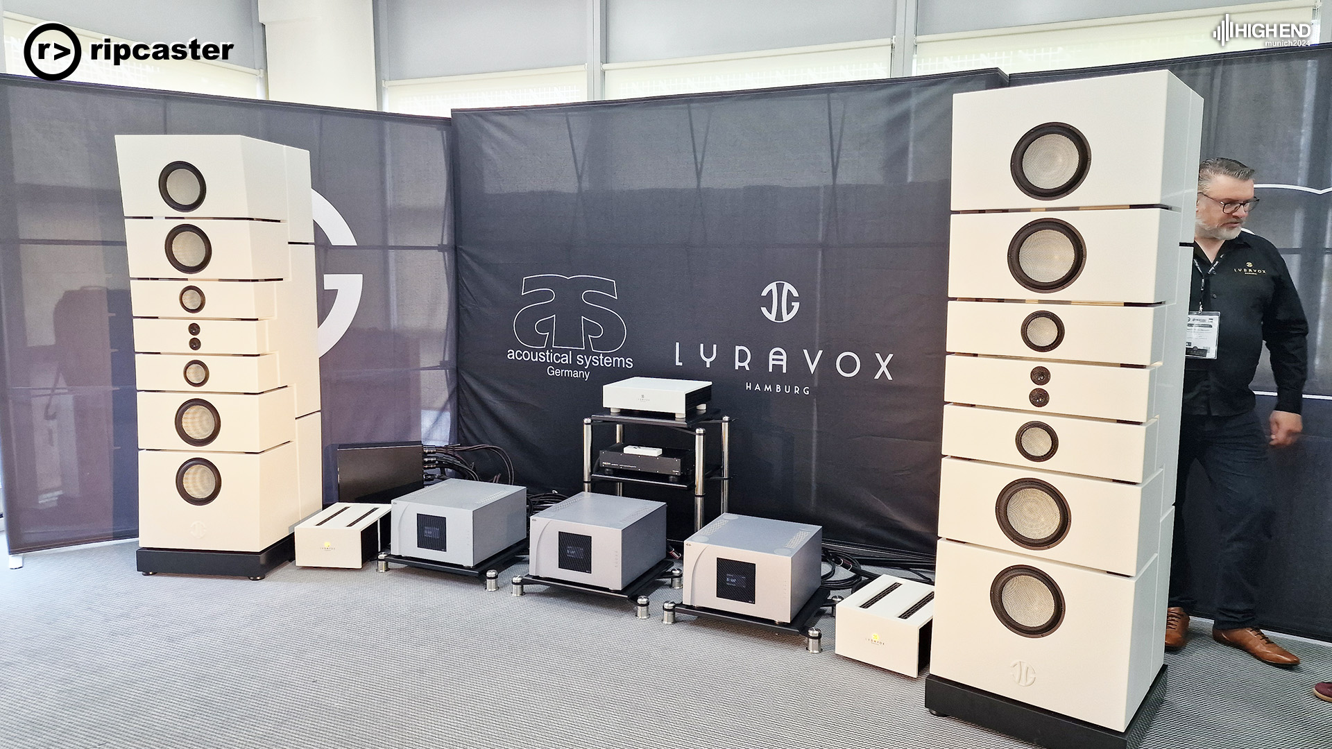 A pair of large white floorstanding speakers with HiFi equipment down on the floor between them.  There's a man emerging from behind one of the speakers.  The speaker is taller than the man