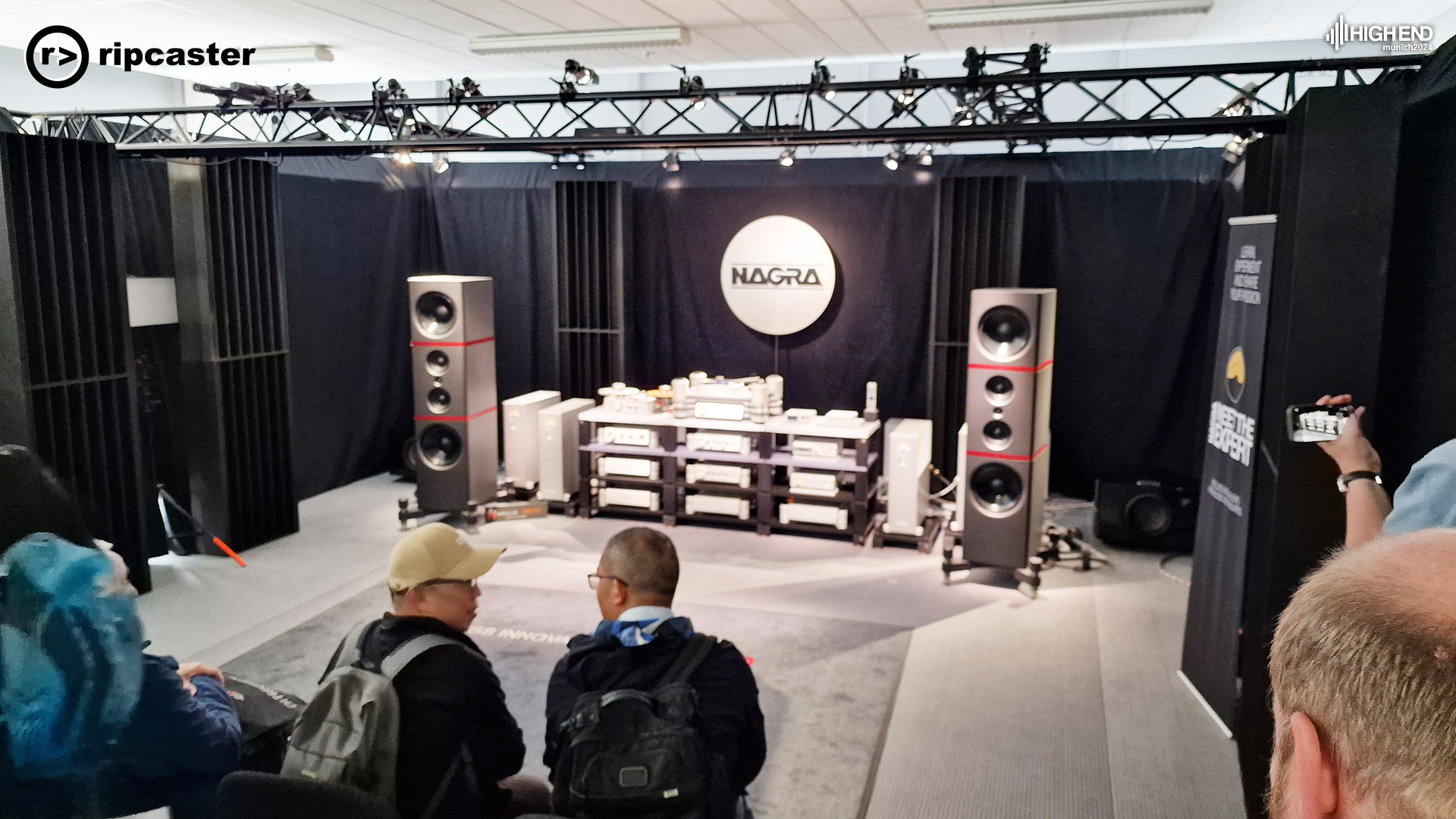 A pair of Nagra speakers with a lot of other HiFi equipment on shelves between them.  there are people in the foreground facing the equipment