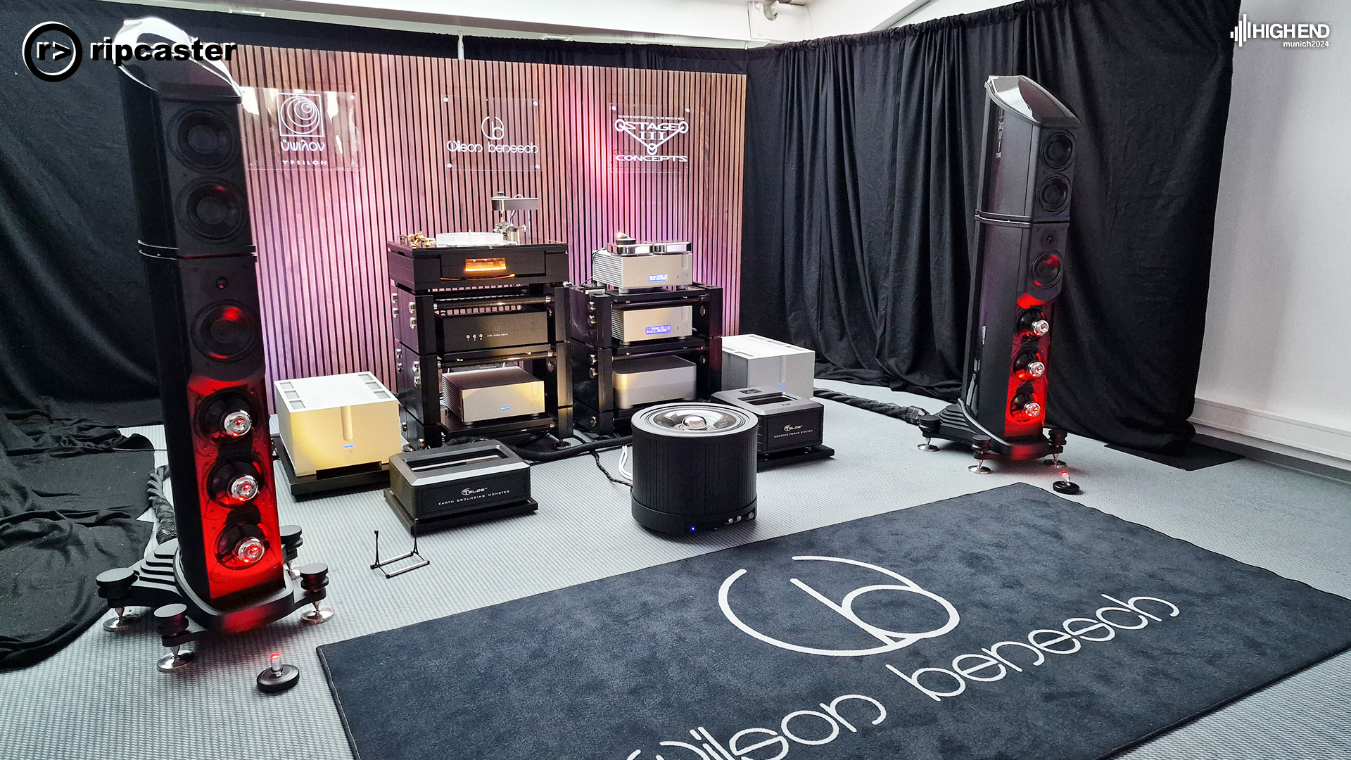 A pair of floorstanding speakers in black with red lights reflecting on them.  Various HiFi equipment is between the speakers