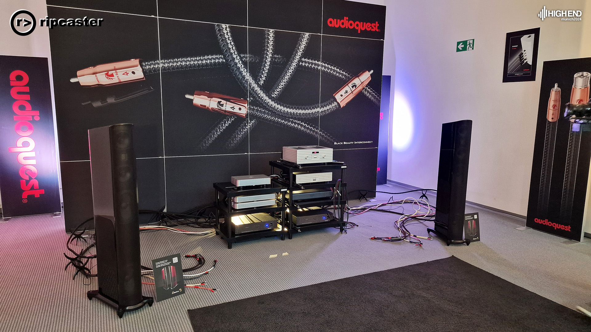 Floor standing speakers in black either side of HiFi equipment.  the backdrop is a large Audioquest one.