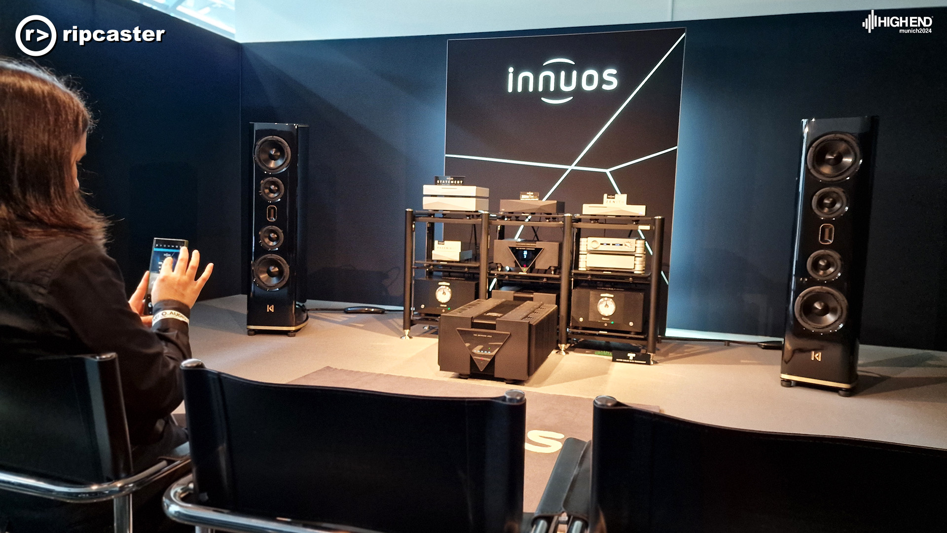 A woman in the foreground to the left on her phone.  In front of her are a pair of black floorstanding speakers with various bits of HiFi kit between them.