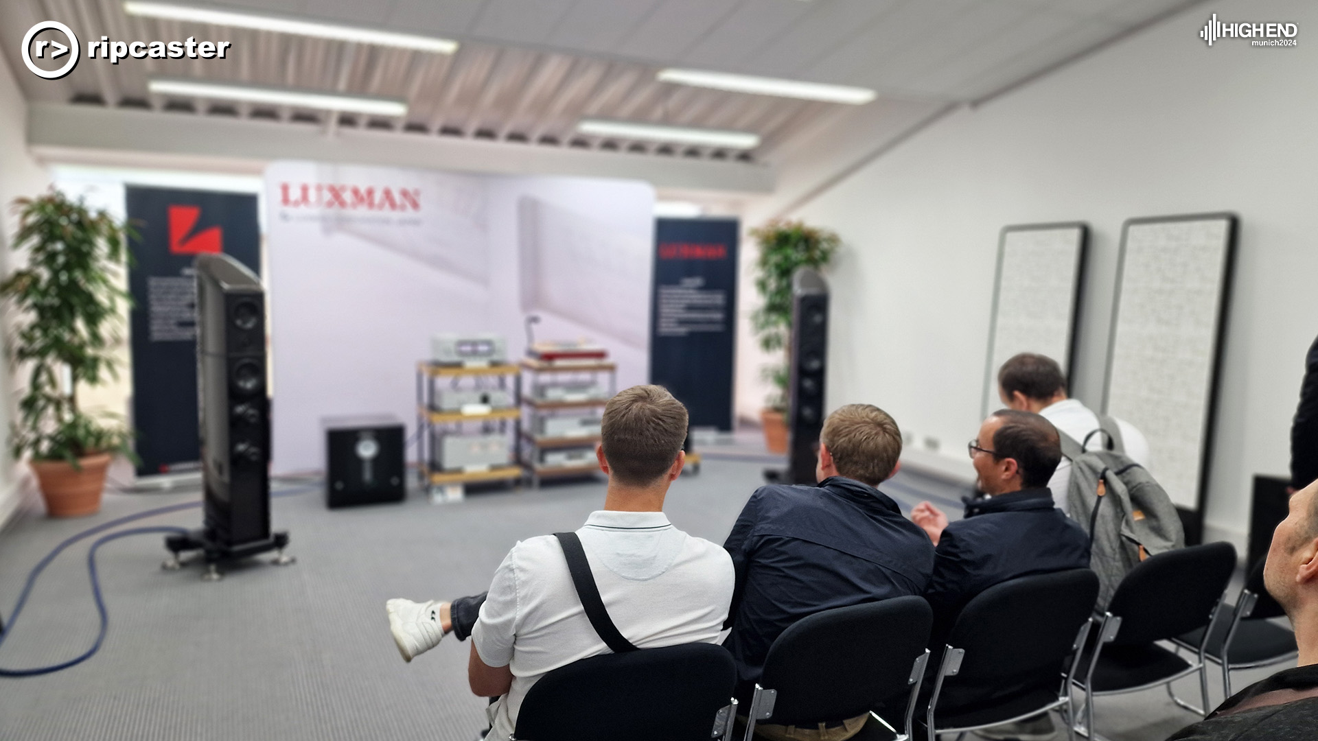 Luxman.  the room with four men in the foreground sitting on chairs.  Speakers and other HiFi kit at the front of the room.