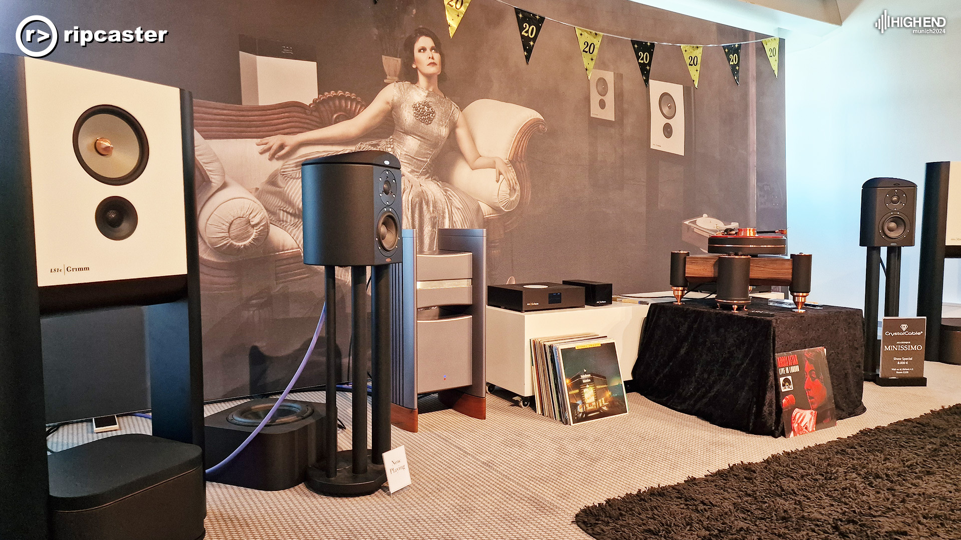 The front of the Grimm Audio room with the same poster of the lady reclining.  There are two pairs of speakers either side of the image.  In between are records propped against a table with various Grimm equipment on it.  there's a record player on a box covered in black cloth.