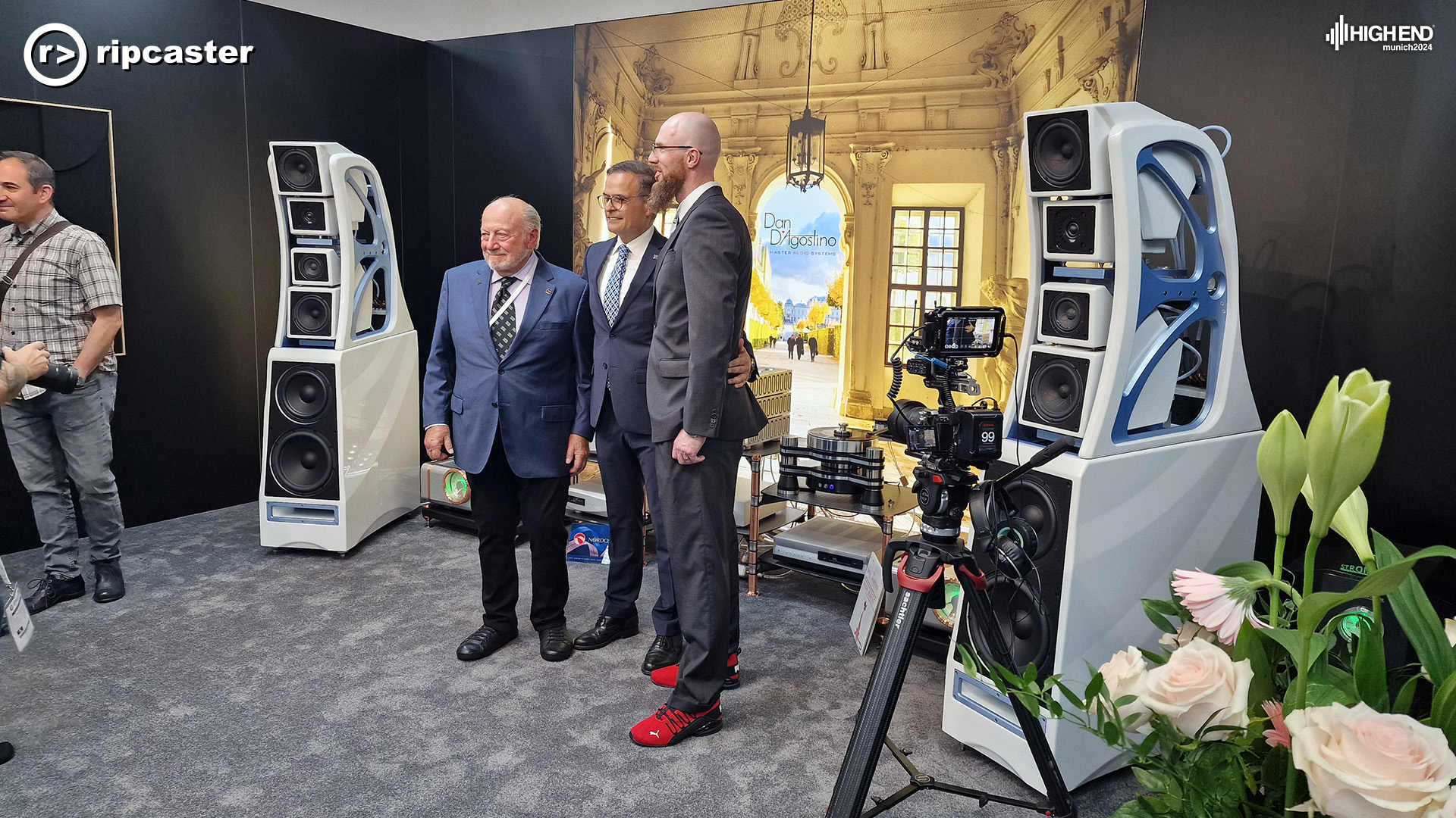 Three men standing between two large white speakers with various pieces of HiFi equipment behind them.