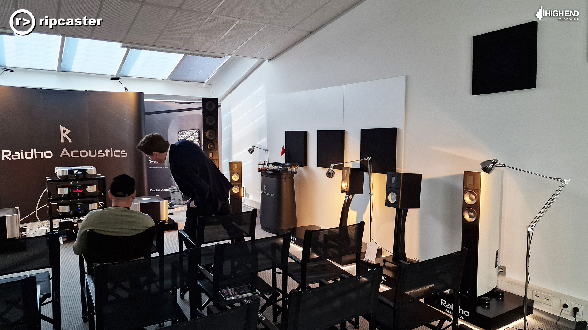 The Raidho Acoustics room showing speakers around the edges and MOON equpment at the front.  There's a man standing leaning to chat to a man sitting