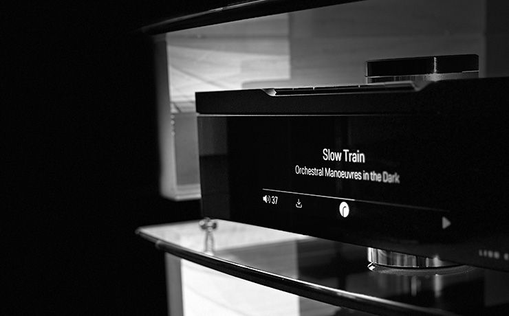 The Linn Klimax DSM on a glass shelf showing the track playing on the screen which is 'Slow Train' by OMD.  The image is black and white