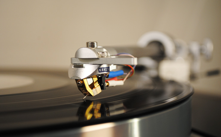 The Krystal cartridge on a tonearm. It's on a record that's spinning. The lighting is warm making the gold shine.