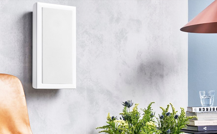 Audiovector On Wall speaker in white with the grille on.  To the left part of a chair is visible.  At the bottom of the image you can see the top of a flower arrangement and to the right is part of a lamp along with a stack of books and a glass of water resting on top of the books