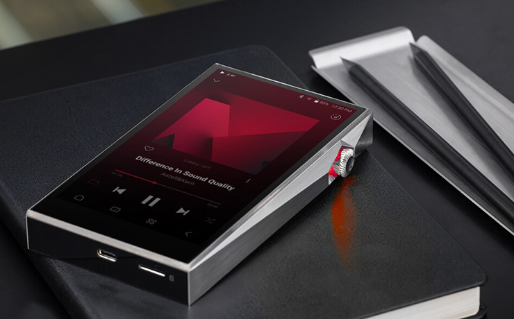 The Astell & Kern SP3000T in silver laying flat on a notebook with two pencils on a wooden tray beside it.