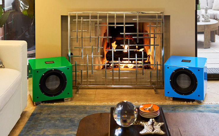 The REL T9x special edition in green on the left and the blue on the right. they are in front of a fireplace with a sofa to the left and objects on a coffee table in the foreground.