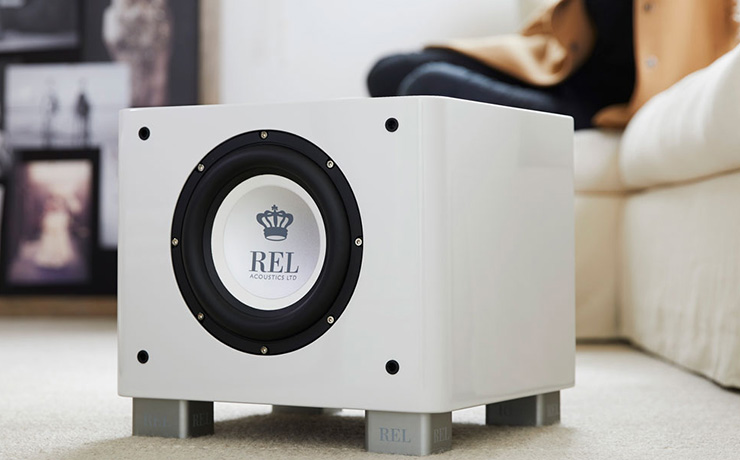 REL T7x subwoofer in white.  on the floor of a living space with a person sitting on a white sofa in the background