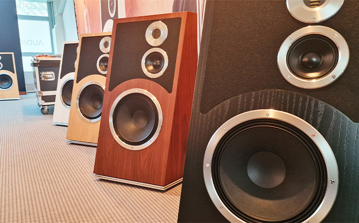 Five Audiovector Trapeze speakers lined up at the HiFi Show in Munich.  Showing the four different finishes.  Black at the front, walnut to the left and back, oak to the left and back and white to the left and back.  Then one Oak one at the back.  the carpet is a light beige with darker beige stripes