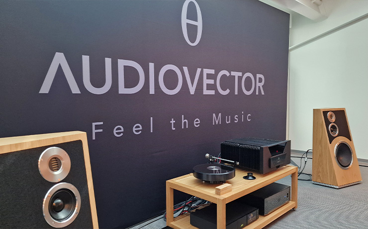 A pair of Audiovector Trapeze speakers in oak either side of some other HiFi equipment on a low unit.  there's a large Audiovector board behind.  This photo was taken at the Munich HiFi Show in 2024