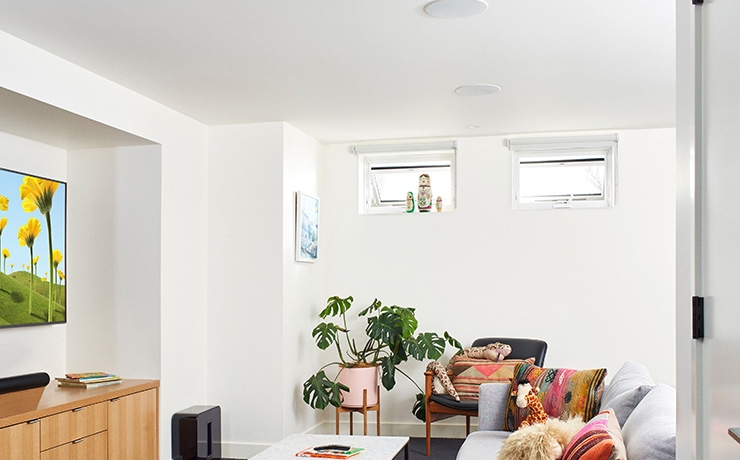 Sonos ceiling speakers in white in a modern living space with a grey sofa, colourful cushions, a plant and a tv on the wall