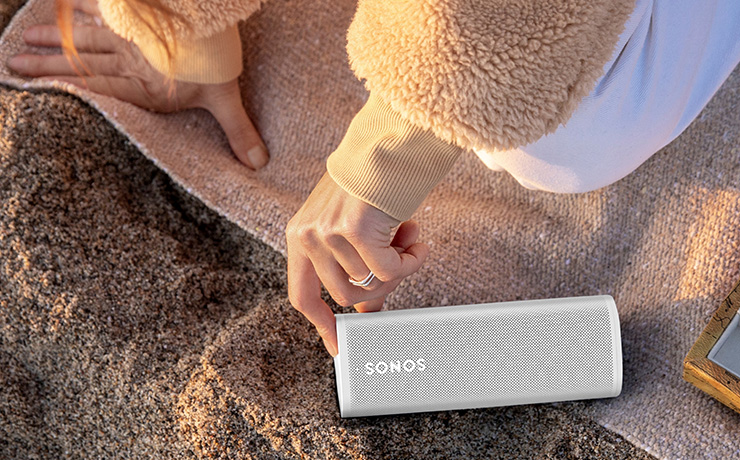 Sonos Roam SL speaker in white on the beach with a person's finger on the control button