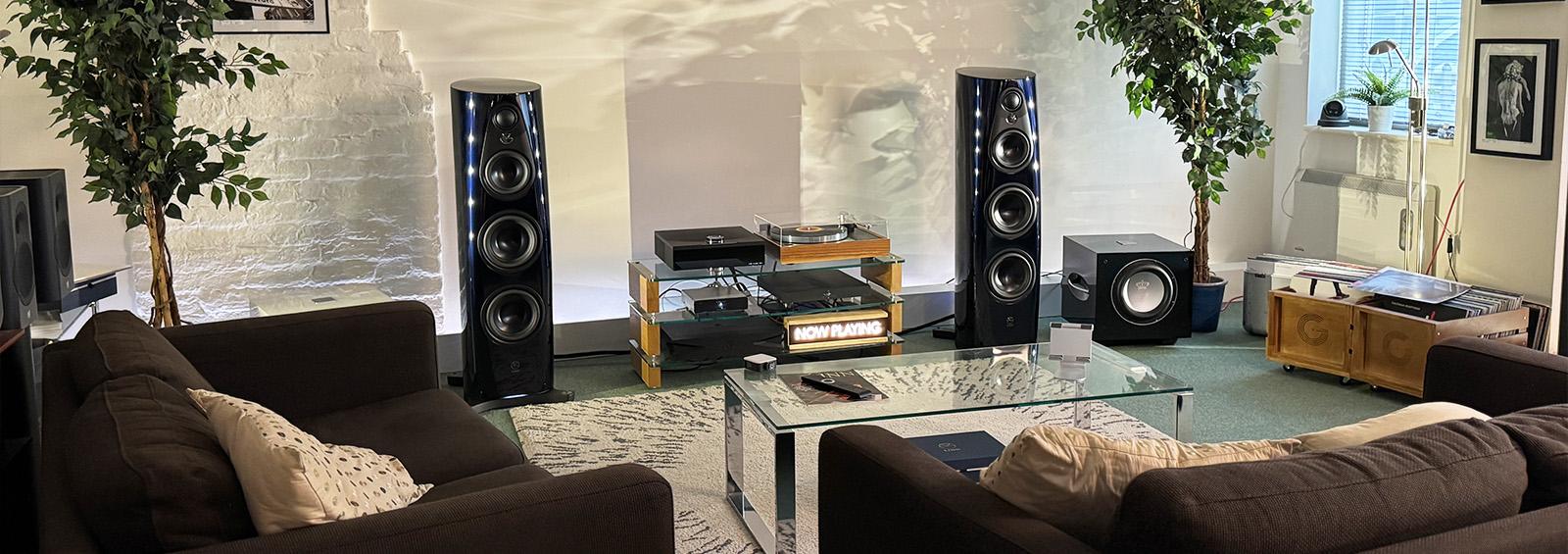 Modern hi-fi listening room featuring a pair of Linn 360 floorstanding speakers, glass equipment rack with a turntable and digital sources, flanked by two large subwoofers and leafy plants. Brown sofas, a glass coffee table with a magazine, remote control, and soft lighting create a relaxed, homely atmosphere.