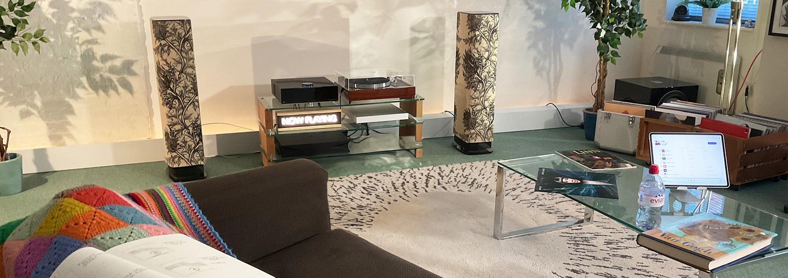 A pair of floorstanding speakers featuring a thistle pattern either side of a HiFi unit with a turntable and a DSM.  There's a sofa in the foreground with a colourful blanket draped over the back which has a notebook resting on top of it.  On the right, there's a glass coffee table with an iPad on a stand on top of it along with an album, bottle of Evian and a Van Gogh book.  There are crates of vinyl on the right behind the coffee table.