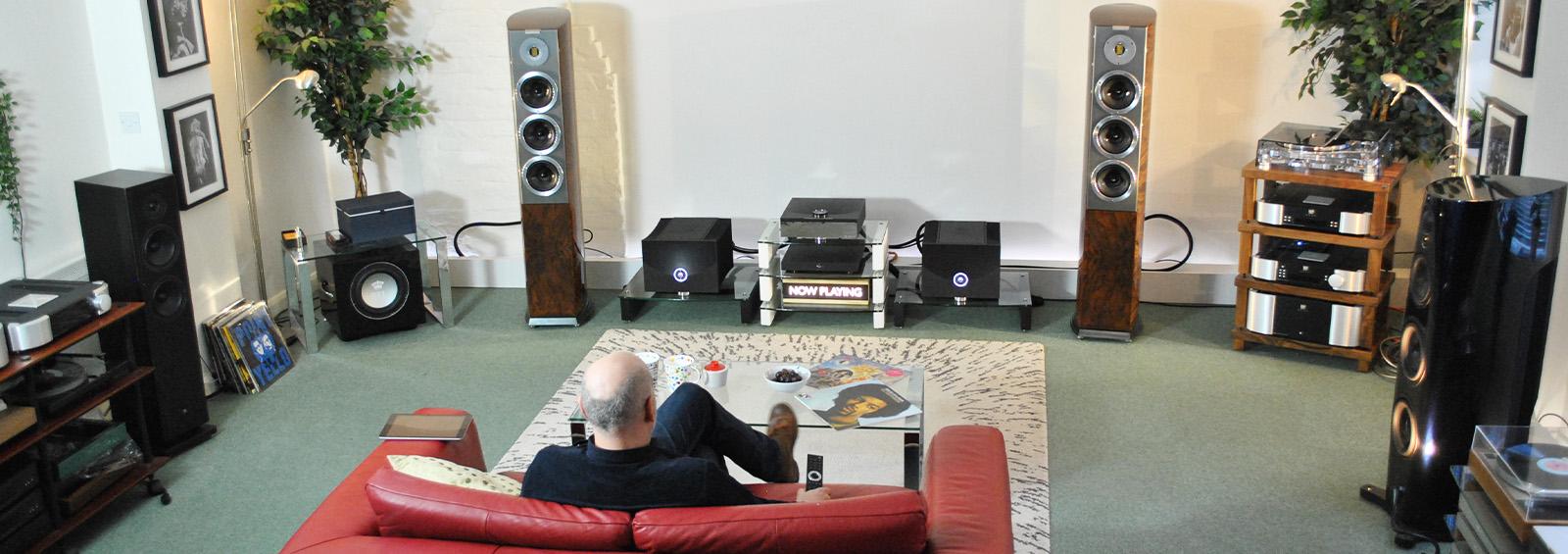 A man sat on a red sofa with his back to us.  In front of him is a glass coffee table and beyond that a HiFi set-up consisting of a pair of Audiovector floorstanding speakers with HiFi kit in between that includes a pair of Linn Klimax 800 Amps and a Linn DSM