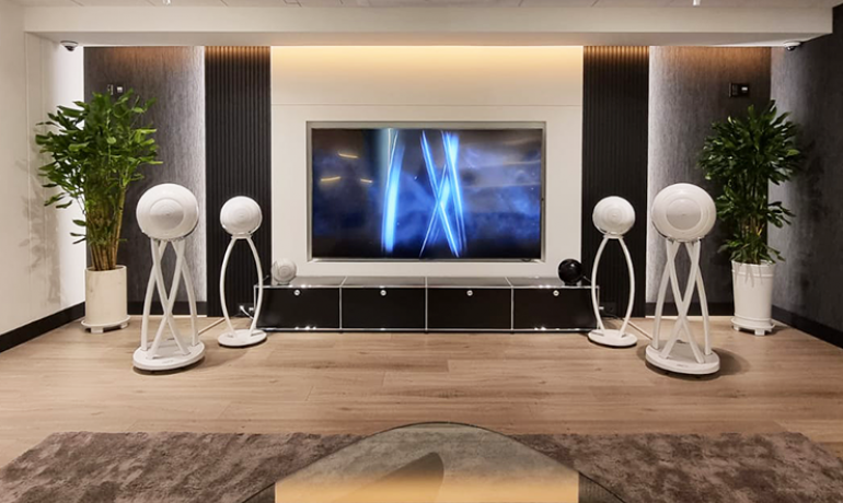 A pair of Cabasse Pelegrina speakers in white either side of a wall mounted flatscreen tv.  Behind them are a pair of Cabasse Pearl speakers in white on stands.  The speakers are in a large living area with a wooden floor and two tall indoor plants in the corners of the room.