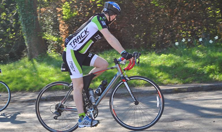 A cyclist on a road with grass and trees behind him.  He is viewed side-on heading for the right side of the photo.  He's wearing cycling clothes in black, white and green.  He is facing away from the camera