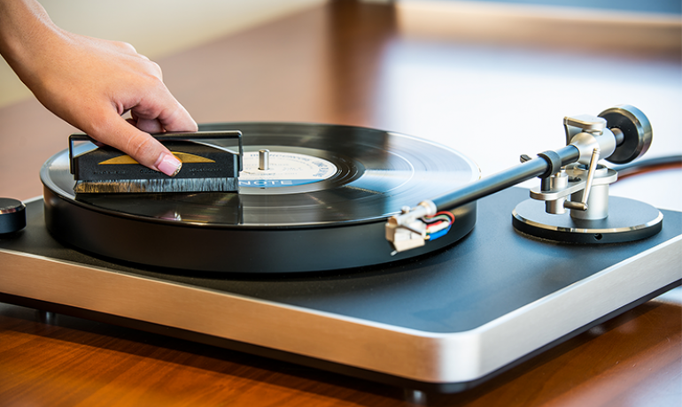 a woman's hand on the left of the image is holding the AudioQuest Super-Conductive Anti-Static Record Brush on a record on a turntable.  The turntable is on a shiny wooden table.