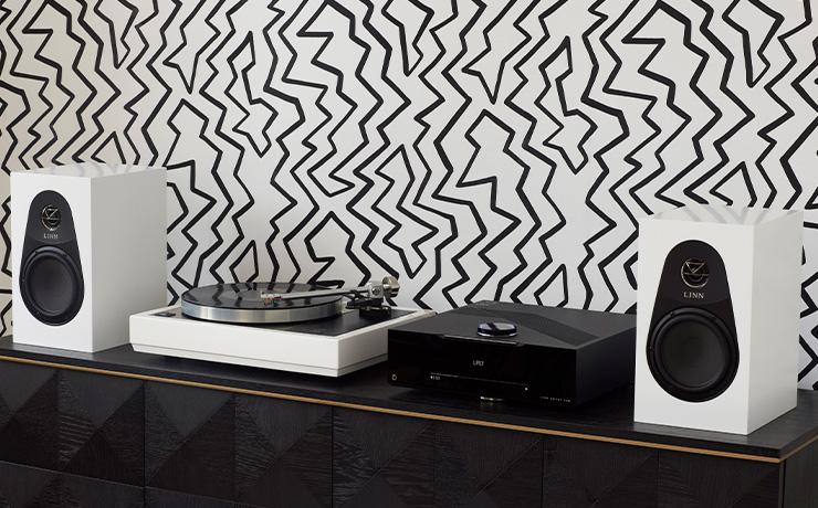 A pair of Linn 119 speakers in white either side of a turntable and a DSM.  The wallpaper in the background is a black and white pattern