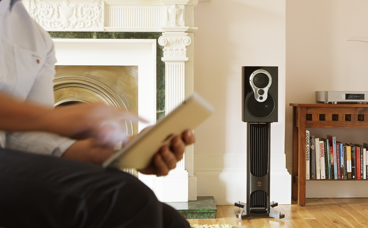 Linn Akudorik Exakt loudspeaker in a living room beside a fireplace with a man in the foreground holding an iPad.