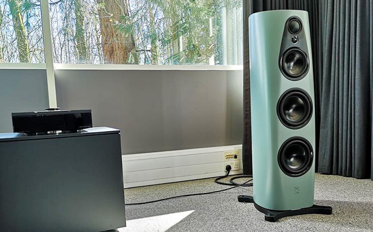 A Linn 360 speaker in a pale blue colour in front of a large window.  There's also a Linn DSM on a unit beside it.