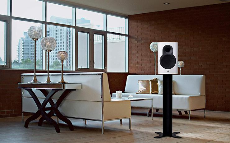 A Kii Seven speaker in white.  It's in a living space with a wooden floor and two cream sofas