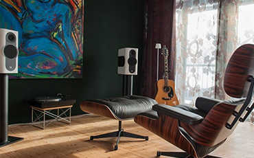 Kii Three speakers in white within a living room setting with a guitar propped in the corner, a large piece of abstract art on the wall, a turntable between the two speakers and a wood and leather easy chair in the foreground