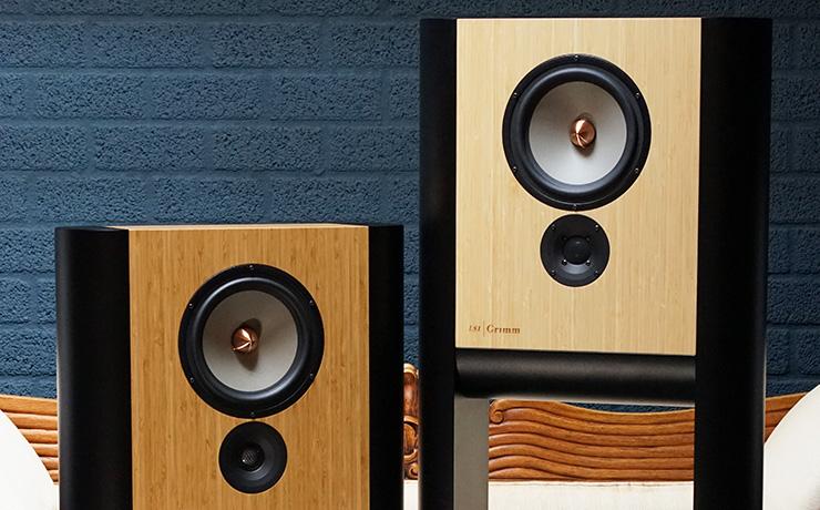 Grimm Audio LS1v2 speakers in two different colours in front of a dark blue brick wall