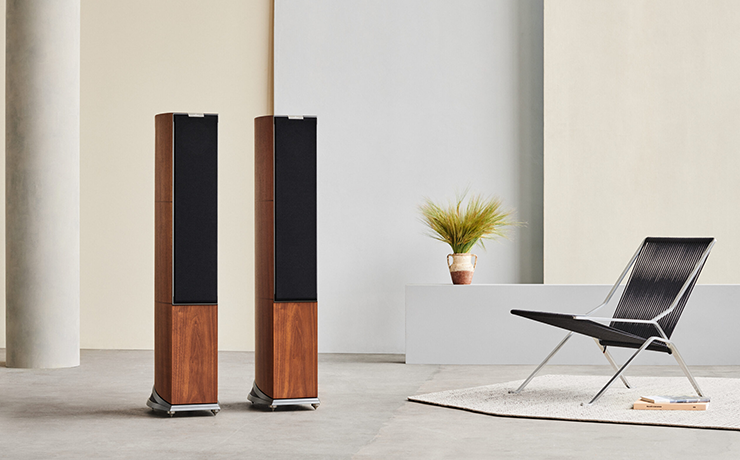 A pair of Audiovector R6 Arreté Loudspeakers with grilles on.  There's a black chair on the right and some long grass on a ledge in the background.