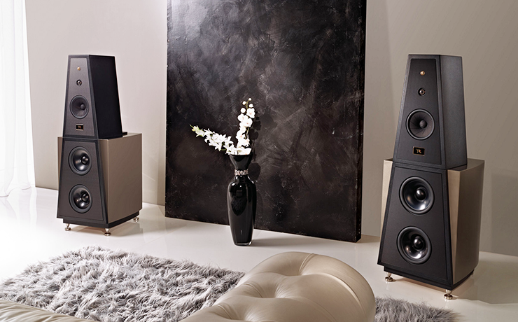 A pair of Rosso Fiorentino Siena Loudspeakers in a living room with a grey rug and beige sofa in the foreground.  Between the speakers there's a large blackboard with a black vase in front of it.  The vase has white flowers in.