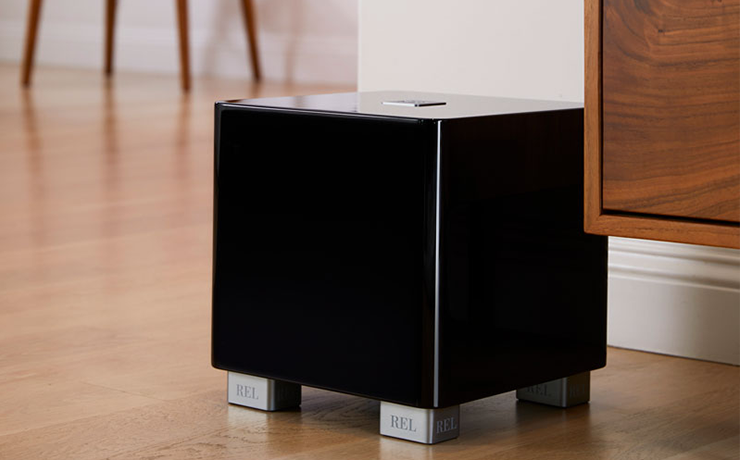 REL T/5x Subwoofer in black on a wooden floor with the legs of a wooden chair in the background and part of a wooden cabinet on the right.