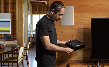 A SONOS in-wall speaker can be seen in the background where a man is holding a Sonos Amp.ng a 
