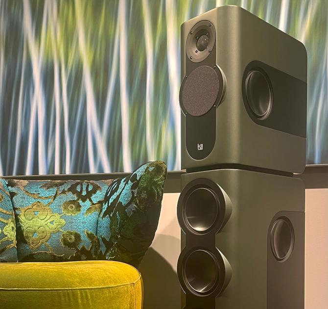 A tall dark green speaker to the right of the image with contemporary art in the background and a chair with a cushion beside the speaker.