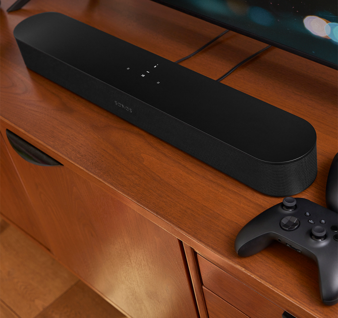 SONOS Beam (Gen 2) on a TV cabinet with a gaming controller beside it.  The bottom of a flat-screen tv is visible.