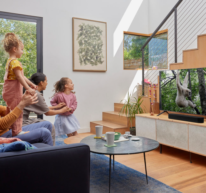 SONOS Beam (Gen 2) in black on a tv cabinet in front of a flat-screen tv which has a monkey on it.  there are stairs to the side of the tv unit.  there's a coffee table in front of the tv unit and a family sitting.  Two children are playing - one is standing up on her dad's legs.  he's seated.