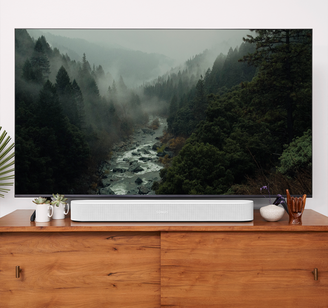 SONOS Beam (Gen 2) in white on top of a wooden tv cabinet.  there's a flat-screen tv showing a picture of a valley with tall trees either side and a rocky river running through.
