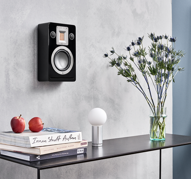 Audiovector QR Wall in piano black on a wall above a table with books and a vase of flowers on.