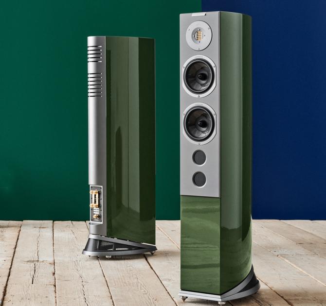 A pair of tall, slim speakers in a green colour on a wooden floor in front of a dark green wall
