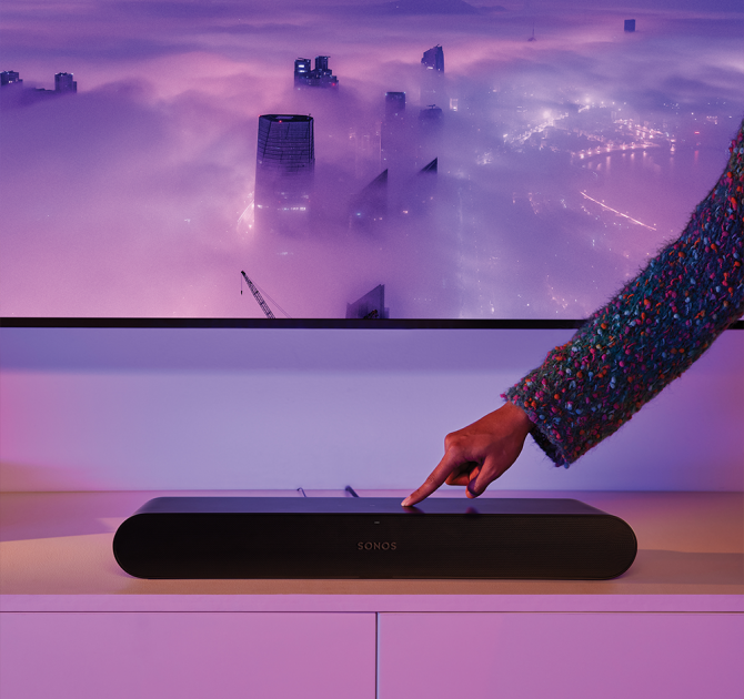 Sonos Ray Smart Soundbar in black in front of a tv with a hand touching the control on the top.