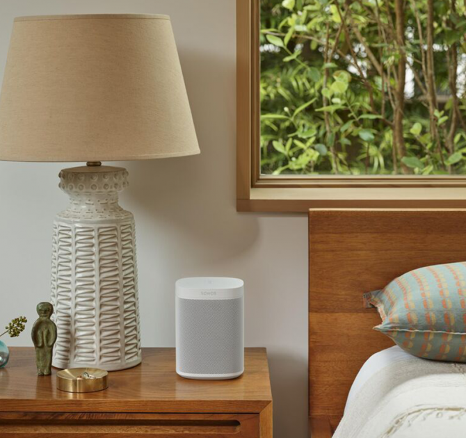 SONOS One SL White standing on a wooden bedside table next to a lamp and a trinket pot with a small statue of a person.  There's a window behind the bed looking out onto the branches and leaves of a bush.