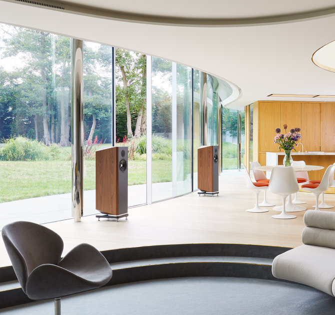 A pair of Kudos Titan 707 speakers with grills off in a large, modern living area in front of a wall of glass doors with a garden beyond.