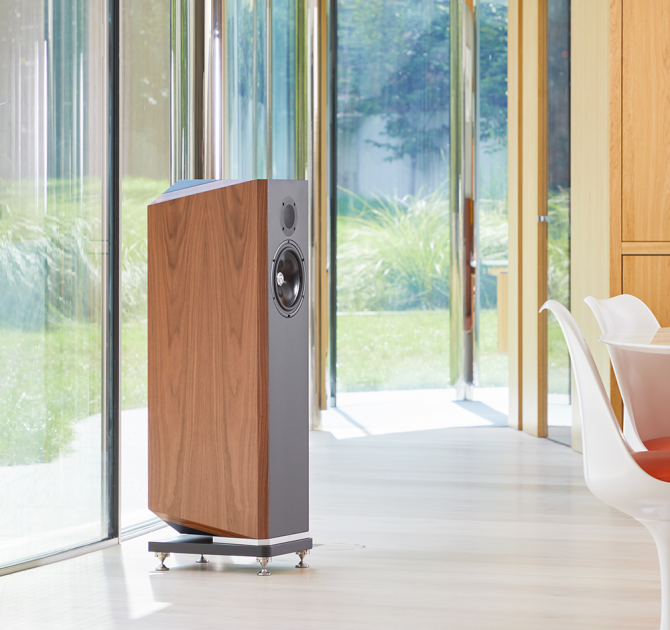 A single Kudos Titan 707 speaker with grills off in a large, modern living area in front of a wall of glass doors with a garden beyond.